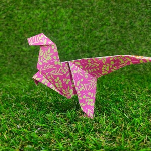 May include: Pink origami dinosaur with a floral pattern on a green grassy surface.