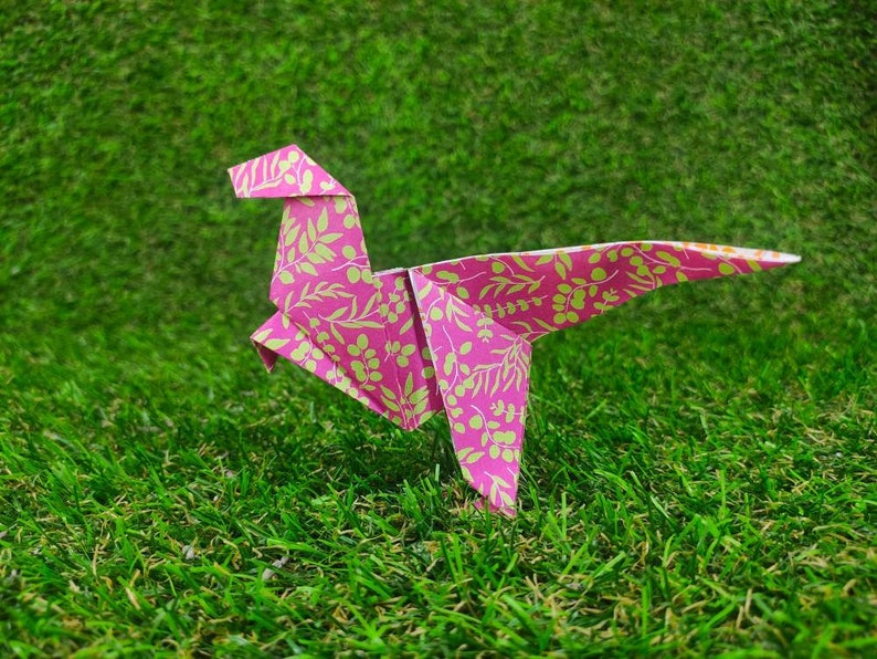 May include: Pink origami dinosaur with a floral pattern on a green grassy surface.
