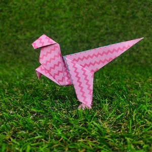 May include: Pink origami dinosaur with a zig-zag pattern, standing on green grass.