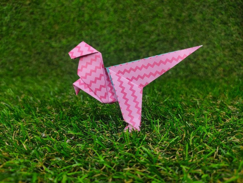 May include: Pink origami dinosaur with a zig-zag pattern, standing on green grass.