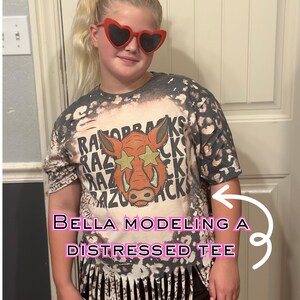 May include: A distressed tee shirt with a graphic of a Razorback hog wearing star-shaped sunglasses. The shirt is a mix of gray and white with a fringed bottom. The text "RAZORBACKS" is printed on the shirt.