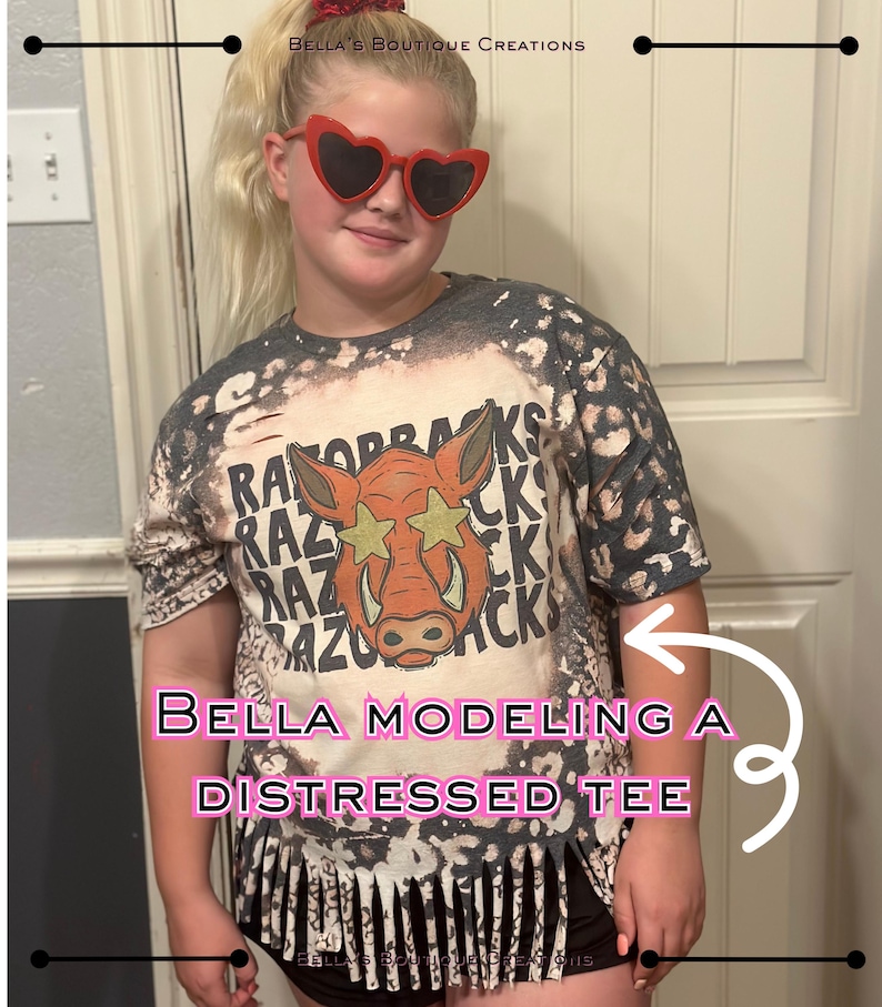 May include: A distressed tee shirt with a graphic of a Razorback hog wearing star-shaped sunglasses. The shirt is a mix of gray and white with a fringed bottom. The text "RAZORBACKS" is printed on the shirt.