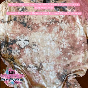 May include: A beige, brown, and black bleached t-shirt with a snowflake pattern. The shirt has text that reads "HANDMADE, TRENDY, ONE-OF-A-KIND BLEACHED TEES & SWEATSHIRTS!" and "UNIQUE BOUTIQUE BLANKS FOR T-SHIRT BARS & MAKERS".