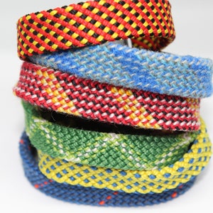 Könnte beinhalten: Ein Stapel aus fünf bunten gewebten Armbändern. Die Armbänder bestehen aus Fäden in verschiedenen Farben und haben verschiedene Muster. Die Armbänder sind übereinander gestapelt, wobei das oberste Armband rot, gelb und schwarz ist.
