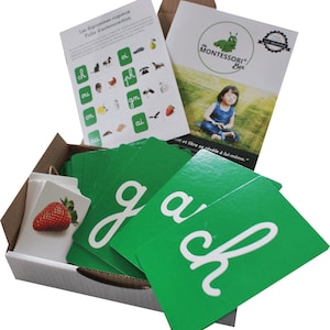 May include: A box of green Montessori letter cards with white letters. The box contains cards with the letters 'g', 'a', and 'ch'. The box also includes a small booklet with the title 'The Montessori Box'.