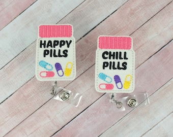 Może przedstawiać: Dwa białe uchwyty na odznaki w kształcie butelki z pigułkami z różowymi pokrywkami. Jeden z nich ma napis "Happy Pills", a drugi "Chill Pills". Oba zawierają kolorowe pigułki.
