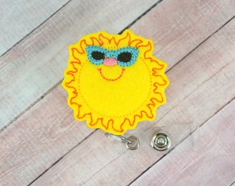 May include: A yellow felt sun with a smiling face and blue sunglasses, attached to a clear plastic badge holder with a silver clip.