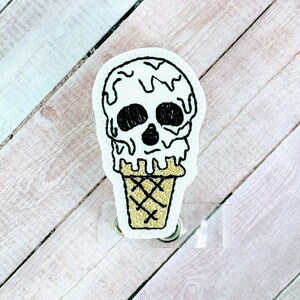 Peut inclure: Un cône de crème glacée en forme de crâne blanc avec un contour noir et de la crème glacée qui coule. Le cône est sur un porte-badge en plastique transparent.