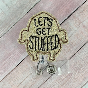 Op de afbeelding: Een gouden glitter badgehouder in de vorm van een gebraden kalkoen met de zwarte tekst "LET'S GET STUFFED" op de voorzijde. De badgehouder is voorzien van een doorzichtig plastic riempje met een metalen drukknoop en een intrekbaar mechanisme.