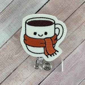 May include: A white embroidered mug badge reel featuring a dark brown coffee fill, a smiling face, and a textured orange scarf wrapped around the center. The design is attached to a clear plastic clip with a circular metal spring mechanism.