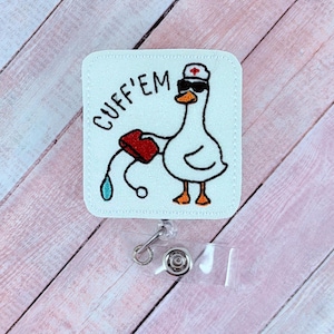 May include: A white badge reel with a cartoon goose wearing sunglasses and a nurse's hat, holding a red container with a blue droplet. The text "CUFF'EM" is written on the badge. The reel has a clear plastic strap and a metal clip.