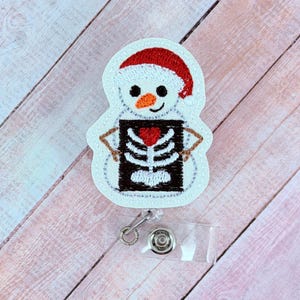 May include: A white snowman-shaped badge reel with a red Santa hat and a black and white skeleton design. The snowman has a carrot nose and a red heart. The badge reel has a clear plastic clip and a metal retractable cord.