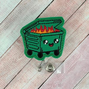 Könnte beinhalten: Ein grünes, gesticktes Ausweis-Jojo mit einem Cartoon-Müllcontainer in Flammen und einem lächelnden Gesicht. Der Müllcontainer hat schwarze Räder und einen leicht geöffneten Deckel. Die Flammen im Inneren sind rot und orange.