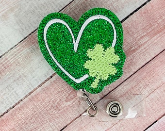 Puede incluir: Portagafete en forma de corazón verde con purpurina, contorno blanco y un trébol de cuatro hojas verde en el centro. El portagafete tiene un clip de plástico transparente.