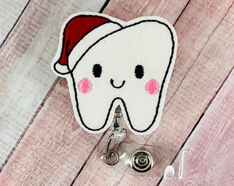 Può includere: Un dente bianco con un cappello di Babbo Natale rosso, sorridente con guance rosa, è attaccato a un porta badge retrattile in plastica trasparente.