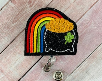 Puede incluir: Un pote de oro negro con purpurina con un arcoíris y un trébol, unido a un carrete de insignia retráctil transparente.