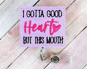 May include: A purple badge holder with the text "I gotta good heart but this mouth" in black with a pink heart. The badge holder has a clear plastic strap and a gold metal clip.