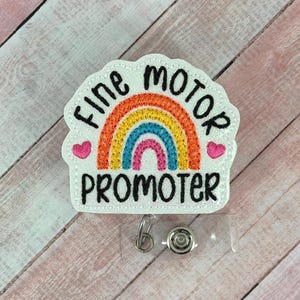 May include: A white badge with the words "Fine Motor Promoter" in black, arched around a rainbow design. The rainbow has layers of red, orange, yellow, blue, and pink. Two pink hearts flank the rainbow. A clear plastic retractable badge reel is attached.
