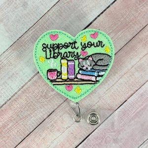May include: A green heart-shaped badge reel with the embroidered phrase "support your library." The design includes a stack of books, a sleeping grey cat, and a pink mug. The reel has a retractable cord and a metal clip.