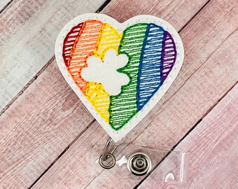 Puede incluir: Un portagafete blanco en forma de corazón con un diseño de arcoíris y una huella de pata blanca en el centro. El portagafete está unido a un carrete retráctil de gafete de plástico transparente.