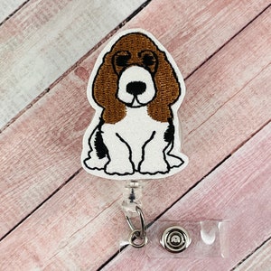 May include: A brown and white embroidered basset hound dog badge reel with a clear plastic badge holder and a silver clip.