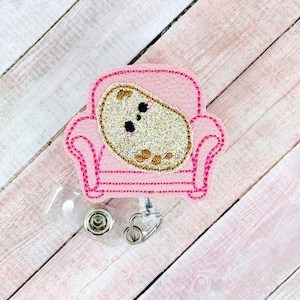 Könnte beinhalten: Ein rosa bestickter Badge-Rollenhalter mit einer Cartoon-Kartoffel, die in einem rosa Sessel sitzt. Der Rollenhalter ist an einem durchsichtigen, ausziehbaren Badge-Halter befestigt.