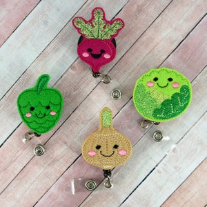 May include: Four embroidered felt vegetable badge reels with smiling faces. The vegetables are a green hop, a brown onion, a green cabbage, and a pink radish.