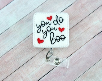 May include: A white square badge reel with the embroidered phrase "you do you boo" in black script, accented by three red hearts. The badge reel has a clear plastic strap and a metal clip.