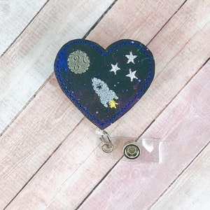 May include: Heart-shaped badge reel featuring a space-themed design. The design includes a rocket ship, moon, and stars against a dark blue and purple galaxy background. The reel has a retractable cord and a clear plastic clip.