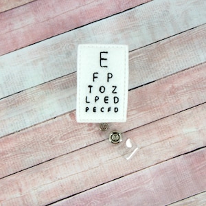 May include: A white felt badge reel featuring a black embroidered eye chart. The chart displays the letters E, F, P, T, O, Z, L, P, E, D, and PECFD. The reel has a clear plastic retractable cord and a metal clip.