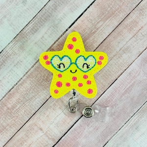 May include: A yellow starfish-shaped badge reel with pink polka dots and heart-shaped glasses. The badge reel has a clear plastic strap and a metal clip for attaching to clothing. The design is whimsical and colorful.