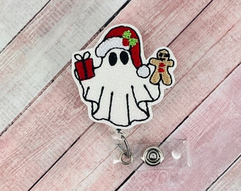 May include: A white ghost wearing a red Santa hat and holding a red gift box and a gingerbread man. The ghost is attached to a clear plastic retractable badge holder.