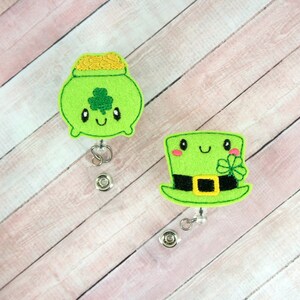 Pode incluir: Dois rolos de crachá de feltro verde com um design de desenho animado bonito. Um rolo apresenta um pote de ouro com um trevo e o outro apresenta um chapéu de duende com um trevo.
