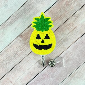 May include: A yellow pineapple-shaped badge reel with a jack-o'-lantern face. The badge reel has a green top and black eyes and mouth. The reel is attached to a clear plastic holder with a metal clip.