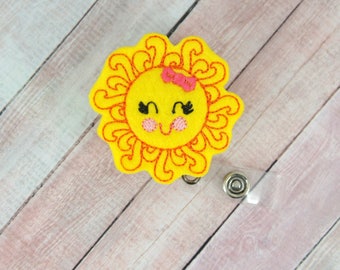 May include: Yellow felt sun with a smiling face, pink bow, and black eyes. The sun is attached to a clear plastic badge holder with a silver metal clip.