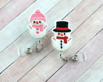 Könnte beinhalten: Zwei Schneemann-förmige ausziehbare Ausweishalter. Ein Schneemann trägt einen rosa Hut und der andere einen schwarzen Zylinder mit rotem Band.