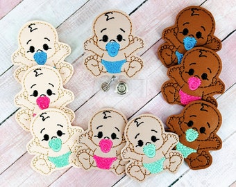 May include: A collection of baby-themed badge reels. The reels feature embroidered baby designs in various skin tones, each with a pacifier and diaper in shades of blue, pink, and teal. The baby designs are beige and brown.