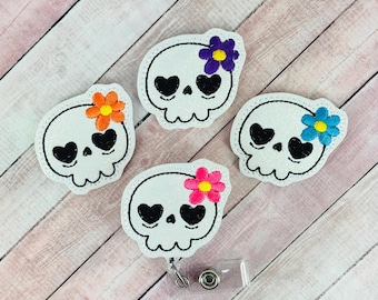Peut inclure: Quatre enrouleurs de badge en forme de crâne blanc avec des yeux en forme de cœur noirs et des accents de fleurs colorées. Les fleurs sont dans des tons d'orange, de rose, de violet et de bleu. Chaque enrouleur de badge a un support en plastique transparent et un clip en métal.