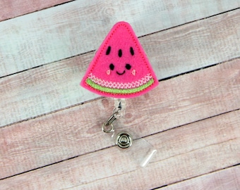 May include: A pink felt watermelon slice with a smiling face and green felt rind attached to a clear plastic retractable badge holder.