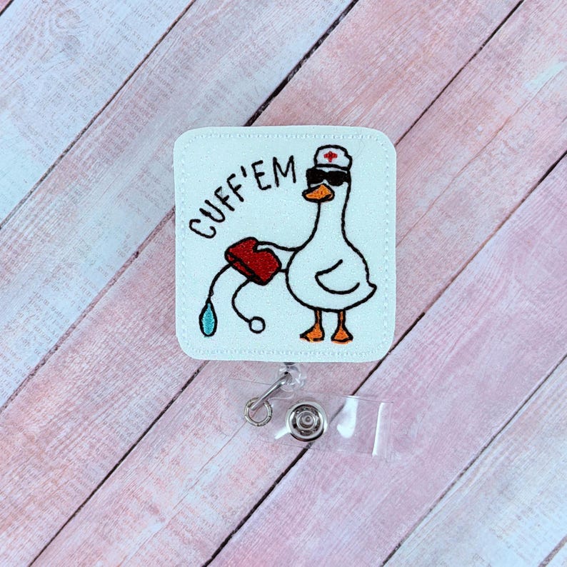 May include: A white badge reel with a cartoon goose wearing sunglasses and a nurse's hat, holding a red container with a blue droplet. The text "CUFF'EM" is written on the badge. The reel has a clear plastic strap and a metal clip.