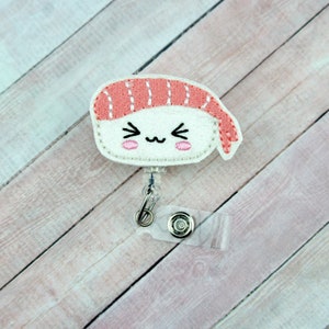 May include: A white and pink felt sushi charm with a cute cartoon face, attached to a clear plastic retractable badge holder.