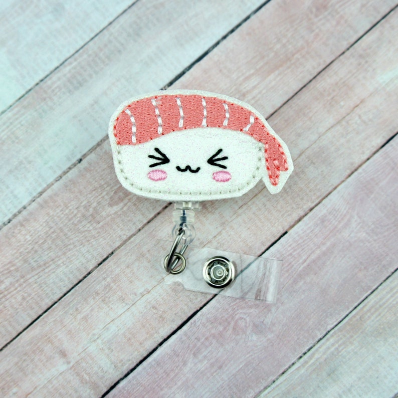May include: A white and pink felt sushi charm with a cute cartoon face, attached to a clear plastic retractable badge holder.