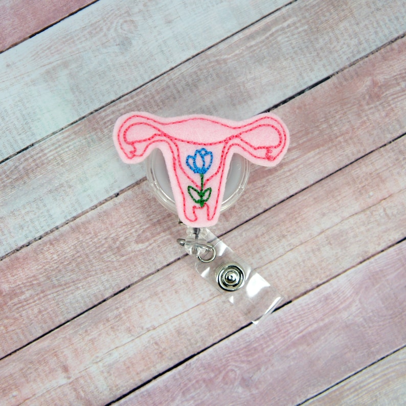 Puede incluir: Un portatarjetas rosa con forma de &uacute;tero con un dise&ntilde;o de flor azul. El portatarjetas tiene una base de pl&aacute;stico transparente y un cord&oacute;n retr&aacute;ctil con un clip met&aacute;lico. El dise&ntilde;o es de fieltro y tiene costuras rojas.