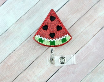 May include: A watermelon slice badge reel with a red glittery surface and black heart-shaped seeds. The badge reel has a green and white rind, and a clear plastic holder with a retractable cord.