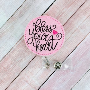 May include: Pink glitter badge holder with a black embroidered saying "bless your heart" and a small pink heart.