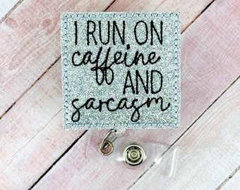 May include: A silver glitter badge holder with a clear plastic badge holder attached. The badge holder has the text "I run on caffeine & sarcasm" in black.