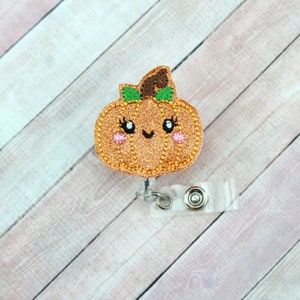 May include: A sparkly orange glitter pumpkin with a green stem and a cute cartoon face. The pumpkin is attached to a clear plastic retractable badge holder with a silver clip.