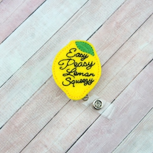 Puede incluir: Portagafete de fieltro amarillo en forma de limón con el texto "Easy Peasy Lemon Squeezy" en letras cursivas negras. El portagafete tiene un carrete retráctil de plástico transparente.