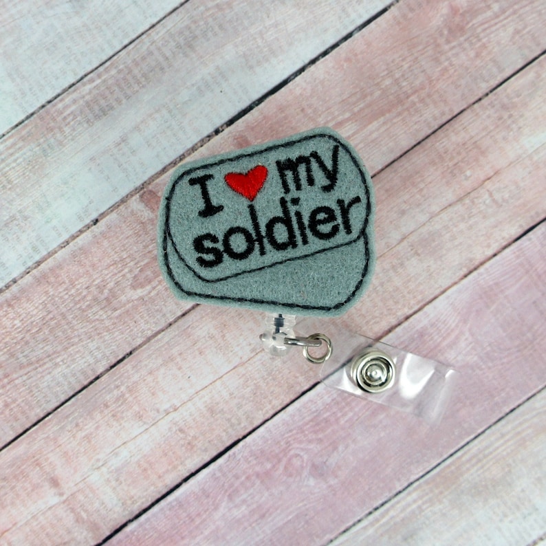 May include: A gray felt badge holder with a red heart and the text "I love my soldier" in black embroidery. The badge holder is attached to a clear plastic retractable badge reel.