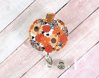 May include: A pumpkin-shaped badge reel with a floral pattern in orange, yellow, and brown. The design includes black spiderwebs and small spiders. The stem is a textured brown fabric. The reel has a clear plastic snap and a metal clip.